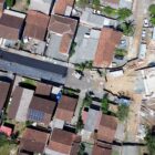 Imagem aérea mostra máquinas trabalhando na pavimentação de uma rua com muitas casas no entorno. Ao fim da rua, está a cabeceira de uma ponte em construção