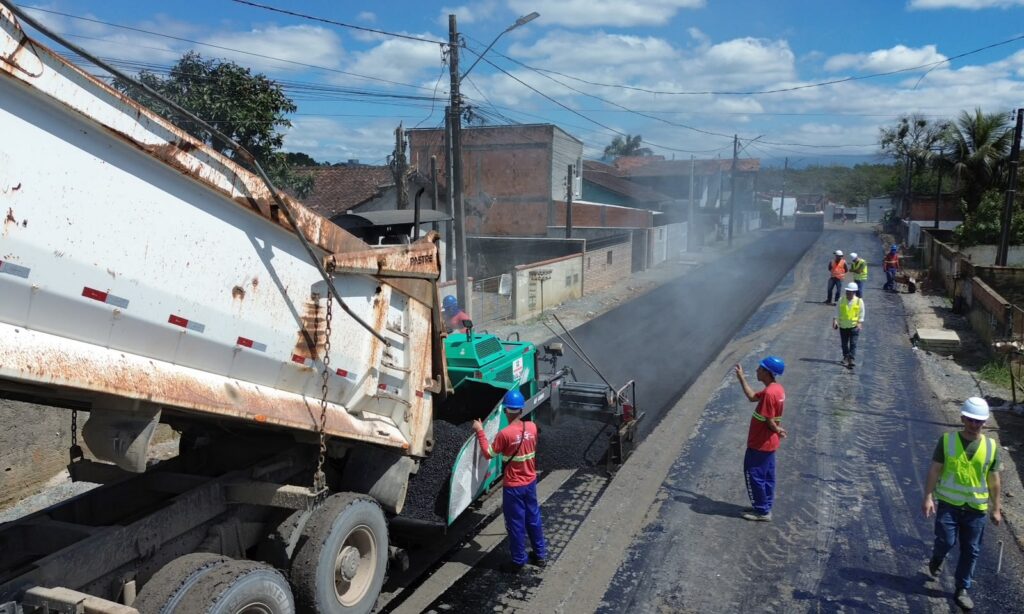 Caminhão e máquina fazem a pavimentação de uma rua com várias casas no entorno. Também há vários trabalhadores uniformizados no local