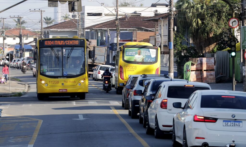 Imagem mostra trânsito intenso em rua pavimentada. Há ônibus, veículos e motos transitando, e fila do lado direito da via