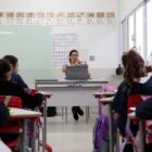 Em uma sala de aula, a professora está sentada na cadeira, com o notebook sobre a mesa, e gesticula para os alunos. Cinco adolescentes aparecem de costas para a câmera