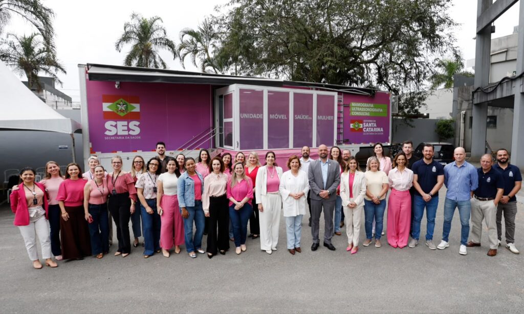Dezenas de homens e mulheres estão em pé, posando para a foto, em frente a um grande conteiner cor de rosa onde funciona o atendimento de saúde da mulher