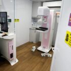 Imagem mostra uma porta aberta com placa dizendo sala de mamografia. No interior, uma mulher de jaleco branco manuseia um equipamento de mamografia