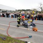 Imagem mostra o simulado de um acidente. Há um jovem caído na pista e cinco bombeiros prestando atendimento a ele, enquanto ao redor dezenas de adolescentes assistem à cena