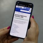 Mãos femininas com unhas pintadas de rosa seguram um celular que mostra o site da Prefeitura, na página sobre o Seminário de Patrimônio