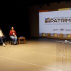 Em um palco, há um homem em pé falando em um microfone e outros dois homens e uma mulher sentados