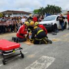 Imagem mostra um simulado de acidente de trânsito em uma rua pavimentada. No local, três bombeiros atendem uma vítima que está deitada no chão. Ao redor, vários adolescentes alunos de uma escola observam