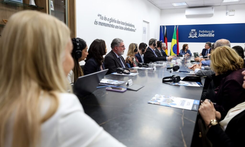 Foto mostra uma sala ampla com uma mesa comprida. Nos dois lados da mesa, há pessoas sentadas em cadeiras, ocupando toda a extensão, em uma reunião.