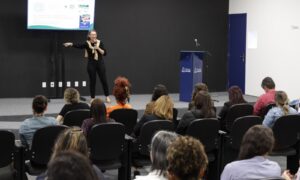 Mulher no palco fala para plateia sentada no auditório