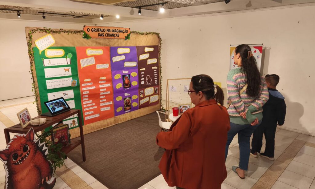 Foto mostra duas mulheres e um menino observando uma exposição escolar, com um grande cartaz e o desenho de um grúfalo