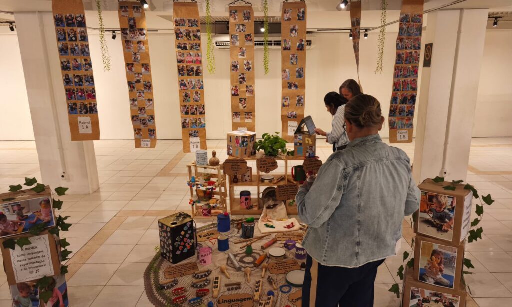 Foto mostra três mulheres observando uma exposição escolar com fotos e trabalhos infantis em um amplo espaço