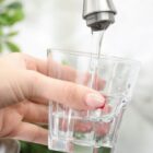 Imagem mostra uma mão feminina segurando um copo de vidro transparente em baixo de uma torneira de inox de onde sai água