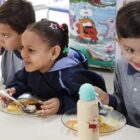 Foto mostra três crianças uniformizadas em uma mesa, cada uma em frente a um prato de comida. Um momento está com talher na mão, a menina no centro sorri e o menino à direita também sorri com o talher na mão.