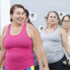 Foto mostra mulher com regata rosa sorrindo em primeiro plano. Atrás dela, há outras três mulheres de diferentes idades em posição semelhante, durante uma aula de funcional