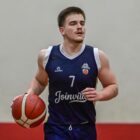 Foto mostra homem branco com regata e calça segurando uma bola de basquete em uma das mãos enquanto anda