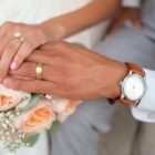 Foto tirada de cima mostra as mãos de uma mulher e de um homem, ambos usando aliança, em primeiro plano. Os dois estão trajados com roupas de noivo e noiva e há um buquê de flores.
