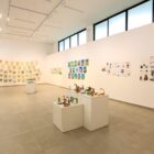 Foto mostra uma ampla galeria de arte com paredes e tanto branco e janelas de vidro. Nas paredes e em alguns blocos no chão, há desenhos e outras obras de arte