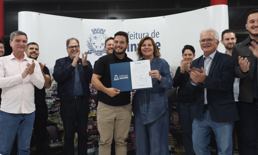 Foto mostra homem e mulher ao centro, rindo e posando para foto enquanto seguram um documento. Ao redor deles, há outros homens e mulheres que aplaudem e sorriem