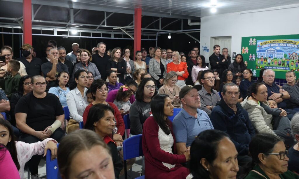 Foto mostra dezenas de homens e mulheres sentados e em pé em um espaço amplo dentro de uma escola
