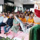 Foto mostra um espaço amplo, com dezenas de pessoas sentadas em cadeiras. À frente, há um personagem feminino de pelúcia em tamanho real