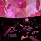 Foto mostra o interior de um domo geodésico com iluminação em tons de rosa. Há uma pessoa deitada em um colchão e outras sentadas.