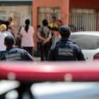 Foto mostra dois homens com uniforme da guarda municipal de costas para a câmera. Eles estão próximos a uma viatura, cuja sirene aparece na foto, perto de uma escola onde há pessoas circulando