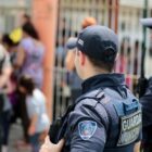 Foto mostra dois guardas municipais de perfil observando a movimentação na saída de uma escola. Ao fundo, há crianças e adultos caminhando