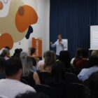 Foto mostra sala ampla com um homem à frente, em pé, falando em um microfone. Na plateia, há homens e mulheres sentados de costas para a câmera