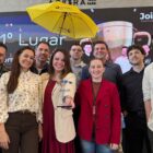 Homens e mulheres estão em pé, sorrindo e posando para a foto. Uma das mulheres à frente segura e exibe um troféu