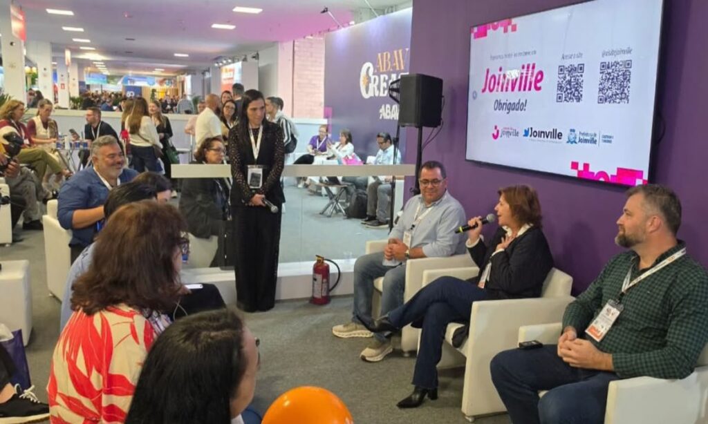 Foto mostra um espaço com três poltronas à frente, com uma mulher sentada no centro falando ao microfone e dois homens. Na frente deles, há homens e mulheres sentados.
