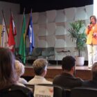 Foto mostra mulher loira falando em um microfone em cima de um palco. À frente dela, na plateia, há pessoas sentadas em cadeiras, de costas para a câmera.
