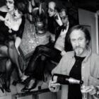 Foto mostra Juarez Machaco, homem branco e magro, segurando uma taça e uma garrafa de champagne. Ele está em pé, em frente a um quadro com uma pintura