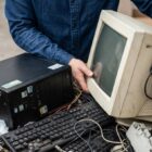 Imagem criada com inteligência artificial mostra os braços de um homem com camisa de manga longa colocando um monitor em uma caixa. Na mesma caixa, também há um CPU, um teclado e cabos