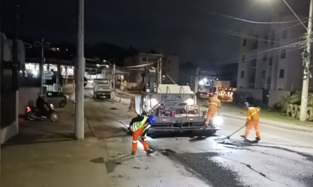 Foto mostra máquina usada na pavimentação e trabalhadores com uniforme laranja em uma rua durante a noite