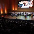 Foto mostra um auditório repleto de pessoas na plateia, de costas para a câmera. No palco, há cerca de 20 jovens de uma orquestra tocando diferentes instrumentos. O telão atrás deles tem a imagem do Natal de Joinville, com uma menina sorrindo