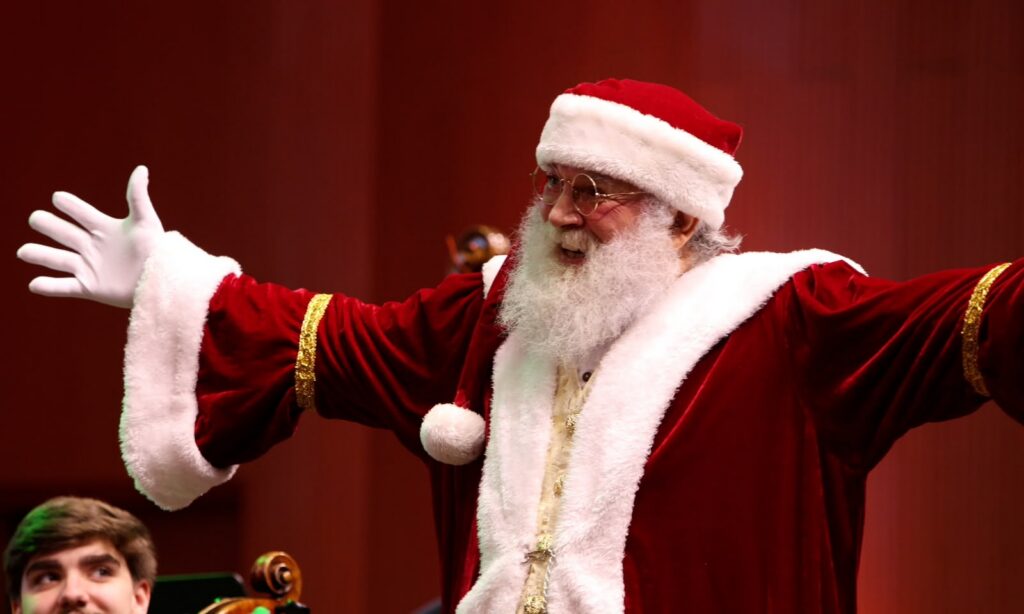 Foto mostra um homem de barba branca e longa vestido com roupa de papai noel. Ele está com os braços abertos e sorri