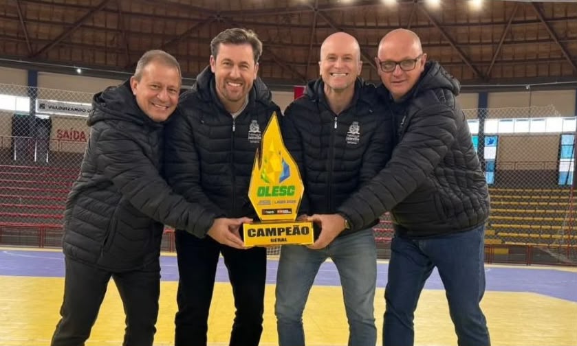 Quatro homens vestidos de preto estão em pé, segurando juntos um troféu em formato de chama que diz "Olesc" e mais abaixo "Campeão"