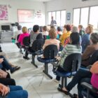 Em uma sala ampla, há dezenas de mulheres sentadas em cadeiras observando um homem em pé, falando à frente da sala