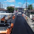 Foto mostra rua sendo pavimentada. Em primeiro plano, há uma máquina com rolo sendo operada por um trabalhador. Ao fundo, há um caminhão despejando o asfalto.