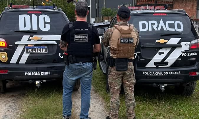 Foto mostra dois policiais civis, um com uniforme preto e outro com uniforme marrom, de costas para a câmera. Eles estão próximos a dois carros da Polícia Civil, plotados com a palavra DIC e DRACO