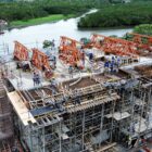 Imagem aérea mostra trabalhadores em uma construção cerca de ferro e concreto próxima a um rio e área de vegetação