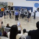 Foto mostra um espaço fechado amplo, em que há pessoas sentadas em uma arquibancada. Um homem e uma mulher estão gravando com o celular a apresentação de uma fanfarra, com adolescentes uniformizados e vários instrumentos