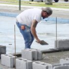 Foto mostra homem usando uma pá no local de uma obra em que está sendo construído um pequeno muro, com tijolos novos dispostos no local