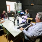 Foto mostra um estúdio de rádio, com paredes com revestimento acústico. Em uma grande mesa, equipada com microfones, estão dois homens e duas mulheres. Eles sorriem