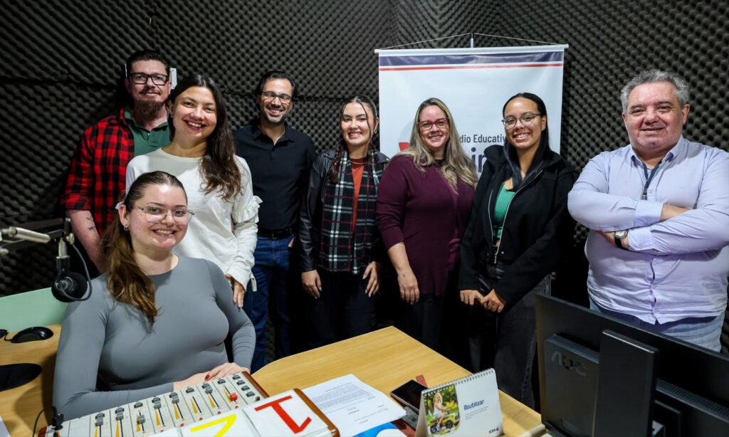 Foto mostra estúdio de rádio com homens e mulheres posando para a foto e sorrindo. Há uma jovem sentada e as demais pessoas em pé