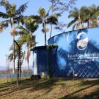 Foto mostra uma área com grama e árvores e uma grande estrutura pintada de azul com a logo da águas de joinville