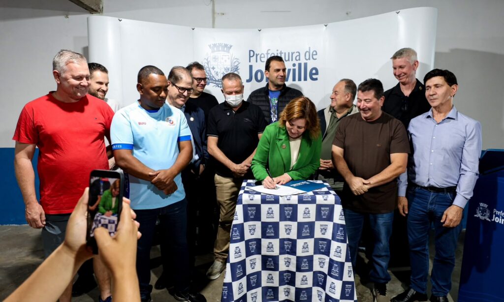 Foto mostra uma mulher e vários homens em pé. Ela está no centro, assinando um documento em cima de uma mesa. Os homens estão ao lado, sorrindo e acompanhando a assinatura