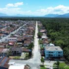 Foto aérea mostra duas ruas paralelas, uma delas se pavimentação, em uma área residencial, com área de mata extensa do lado direito