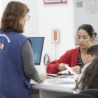 Foto mostra o interior de uma Unidade Básica de Saúde da Família. De costas para a câmera, há uma mulher usando um colete azul, escrito agente comunitário de saúde. Ela está em pé e conversa com uma mulher sentada com uma criança no colo e que manuseia alguns papéis