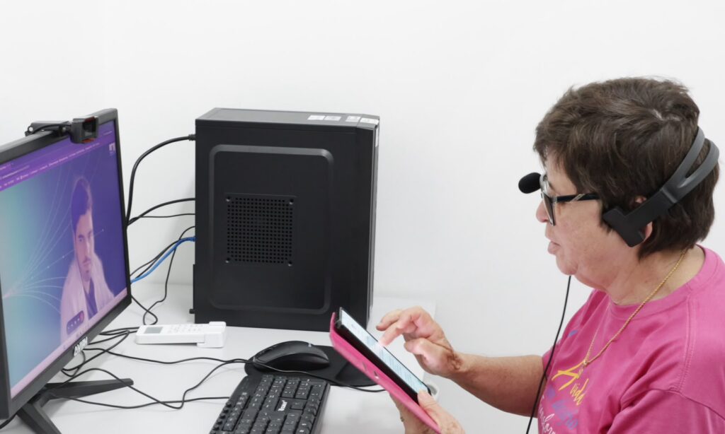 Foto mostra uma videoconsulta. Há uma mulher sentada usando um fone com microfone em frente a um computador. No monitor, aparece um homem de jaleco branco