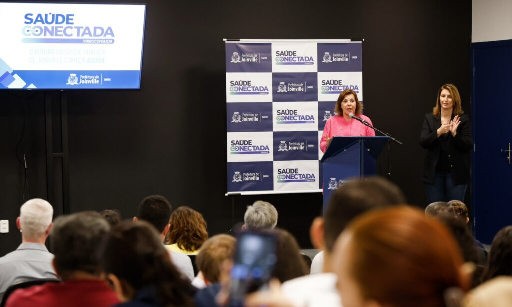 Foto mostra uma mulher loira falando no microfone atrás de um púlpito, em um palco. Ao lado dela, há uma televisão e, atrás, um banner. Na plateia, há dezenas de pessoas sentadas de costas para a câmera
